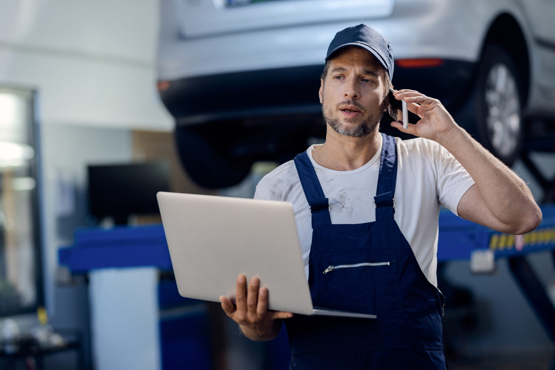 Mid adult mechanic using computer and talking on mobile phone at auto repair shop.